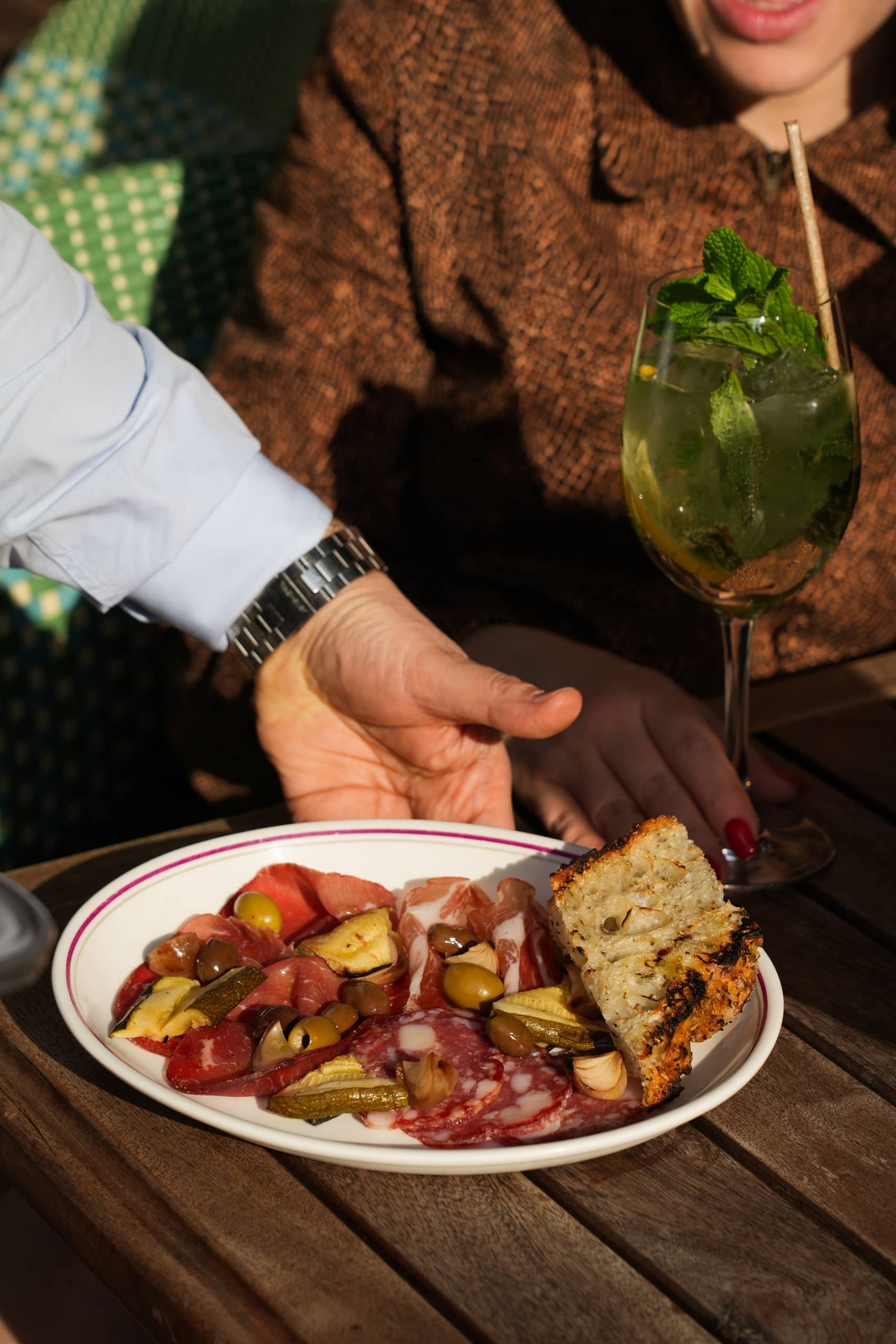 Charcuterie plate with assorted cured meats, olives, pickles, artichokes, and grilled bread served outdoors at Mortimer House Kitchen, paired with a refreshing mint cocktail.