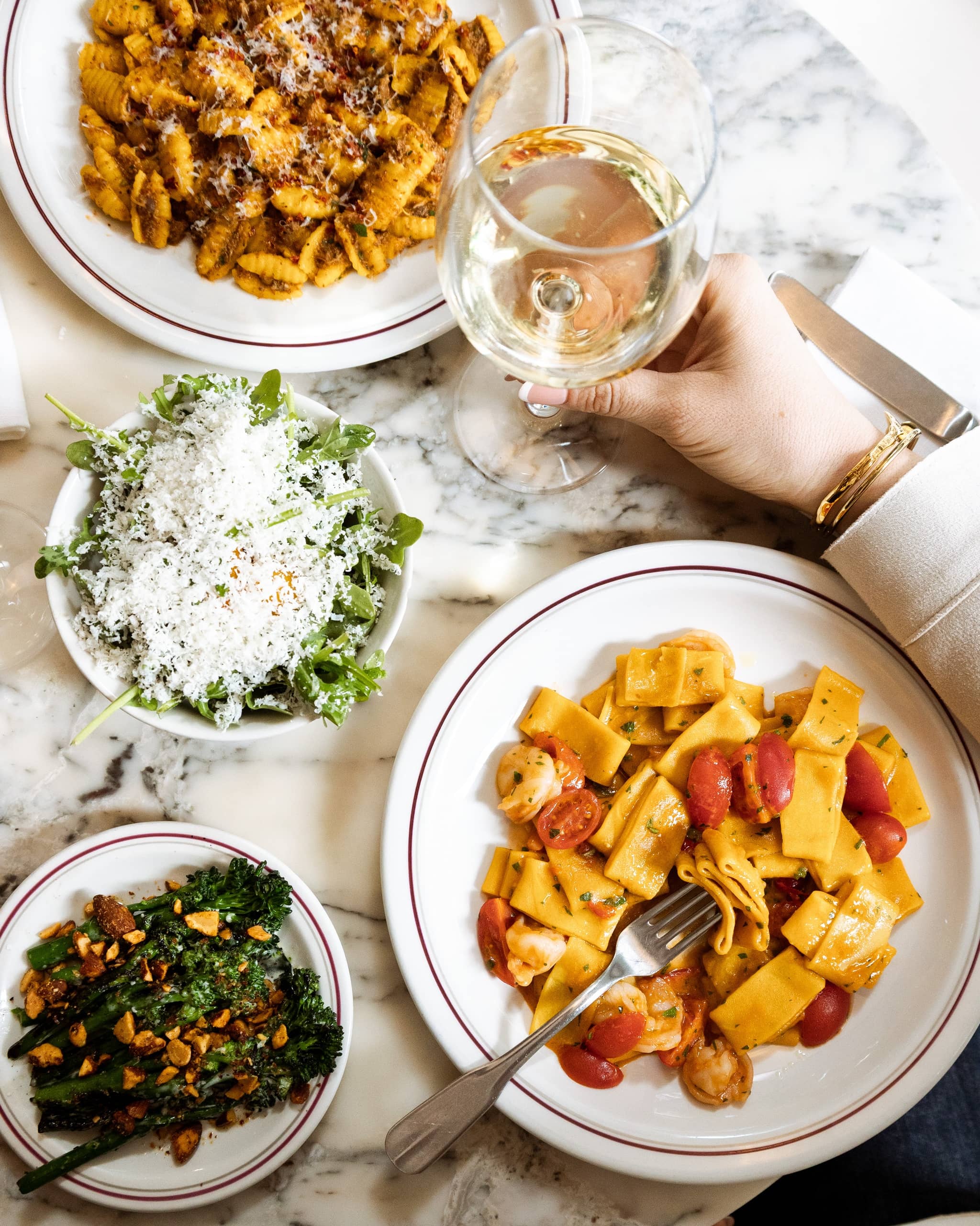 Elegant Italian meal at Mortimer House Kitchen featuring paccheri pasta with prawns and cherry tomatoes, fusilli with rich sauce, arugula salad topped with parmesan, sautéed broccolini with almonds, and a glass of white wine on a marble table.