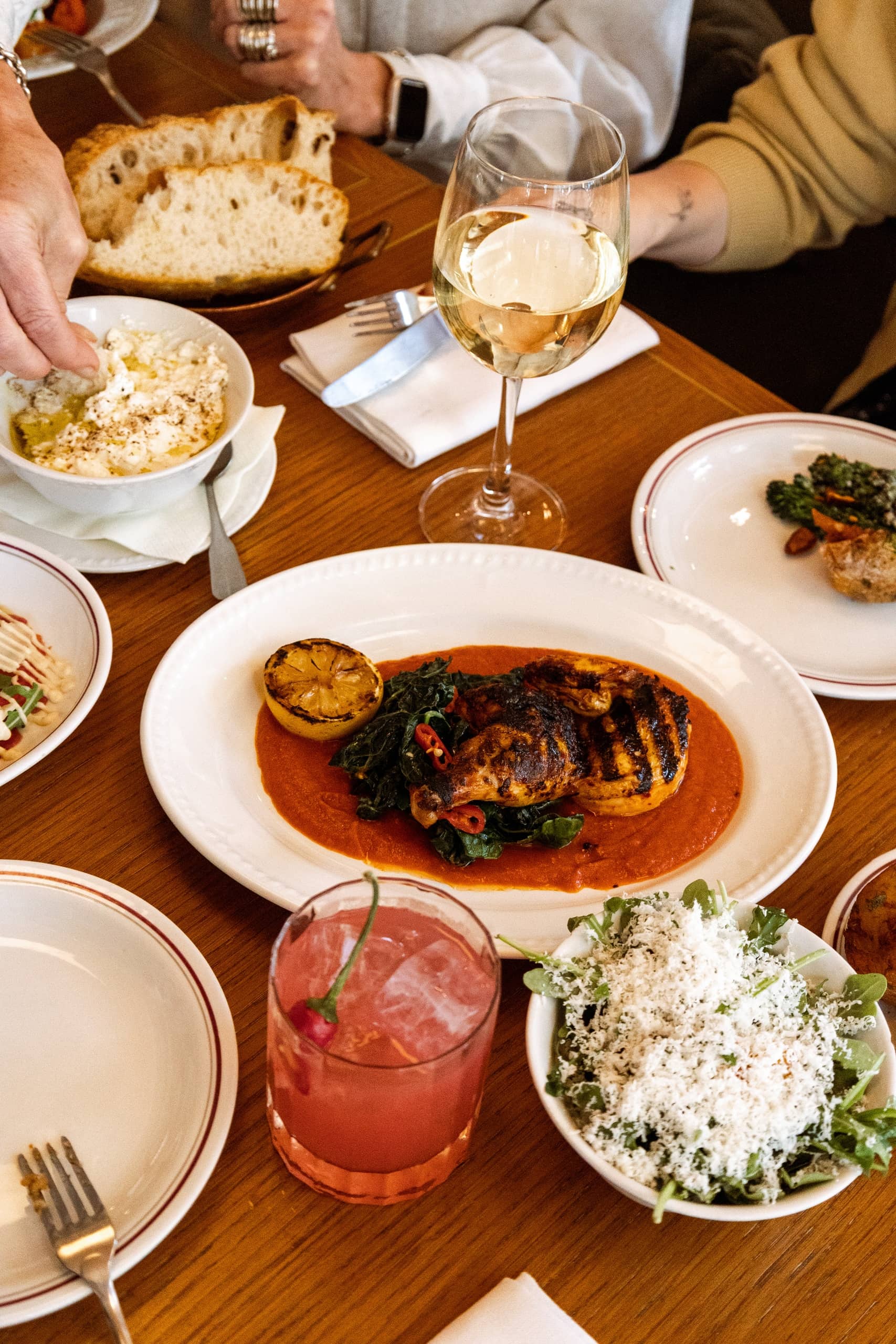 Grilled chicken with wilted greens, charred lemon, and red pepper in a rich tomato sauce, served with fresh bread, burrata, salad, and wine at Mortimer House Kitchen.